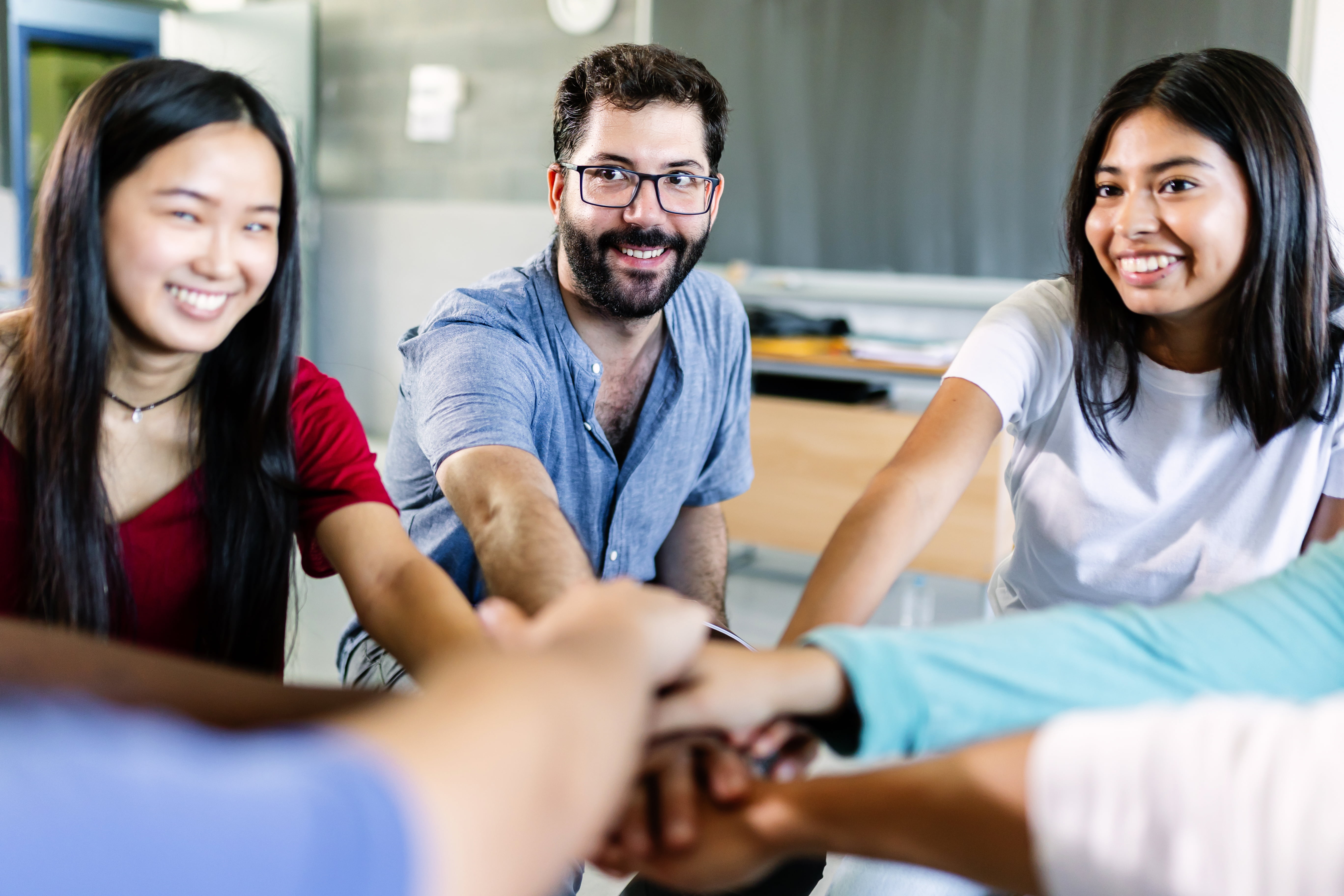 Young multiethnic group of students stacking hands with male teacher in classroom