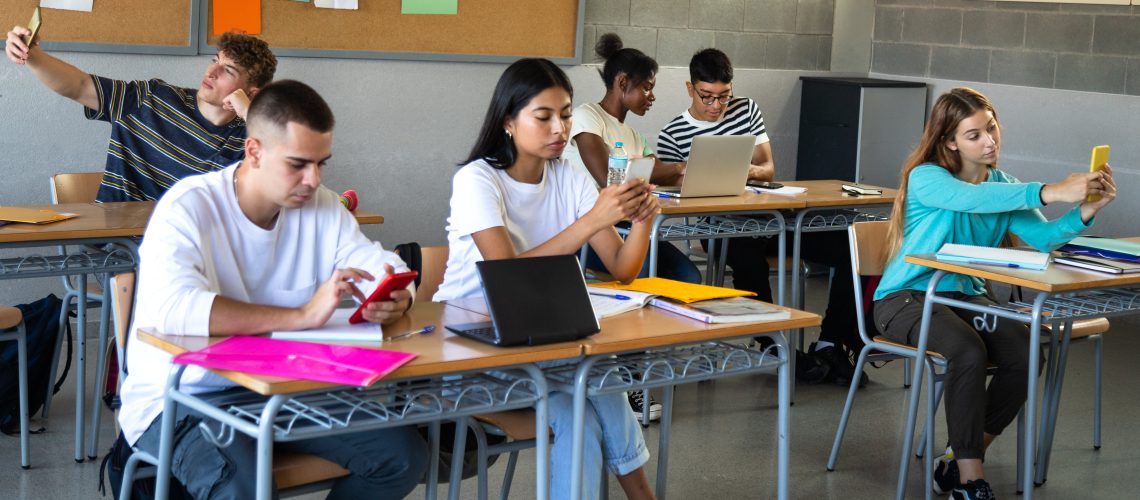 Multiracial high school students distracted from lesson using mobile phones in class.